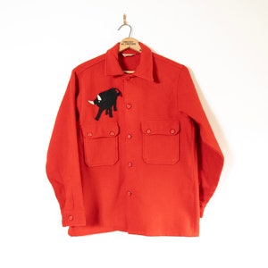 May include: A red long-sleeved shirt with a black bull applique on the front. The shirt has two large pockets on the front and a button-up closure.