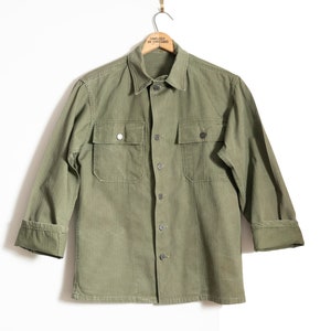 Può includere: Una camicia verde da lavoro a maniche lunghe con due tasche a patta. La camicia ha un colletto abbottonato e una chiusura con bottoni sul davanti.