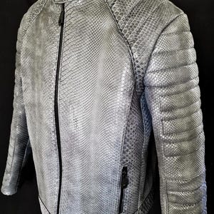 Peut inclure: Veste en cuir grise texturée avec un motif peau de serpent. La veste a un col montant, une fermeture éclair sur toute la longueur et des détails matelassés sur les épaules et les manches. Une petite poche zippée est sur le côté gauche.