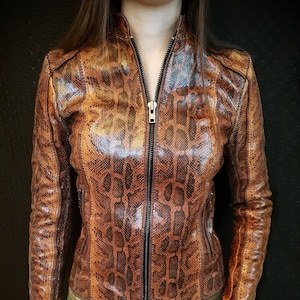 May include: A brown and tan snakeskin-patterned leather jacket with a full-length zip. The jacket has a stand-up collar and is worn with olive green trousers. The jacket's design resembles reptile skin.