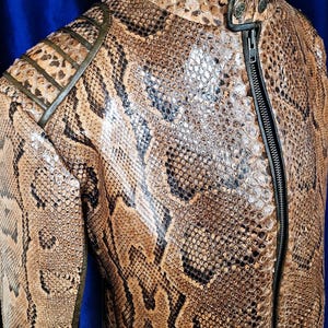 Könnte beinhalten: Eine braun-schwarze Jacke mit Schlangenmuster und schwarzem Reißverschluss. Die Jacke hat einen gerippten Kragen und Schulterakzente. Die Jacke wird auf einer Schaufensterpuppe vor blauem Hintergrund präsentiert.