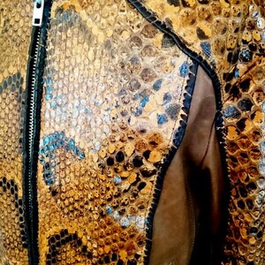 以下が含まれることがあります： ヘビ革模様の茶色とタン色の革製ジャケットのクローズアップ。 ジャケットはジッパー開閉、小さな襟、側面に茶色のスエードパネルが付いています。 ジャケットの襟にはシルバーのボタンが付いています。