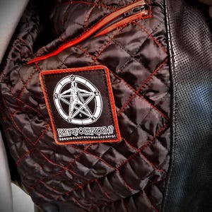 May include: Close-up of the interior of a black leather jacket. The quilted lining is dark brown with red stitching. A rectangular patch with a white pentagram design and the word "PENTAGRAM" is sewn on the lining. A red zipper is visible.