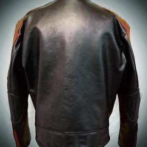 May include: Black leather jacket with orange and red accents on the shoulders and sleeves. The jacket has a classic design with a stand-up collar and a fitted silhouette. The back view shows the jacket's details, including the stitching and panels.
