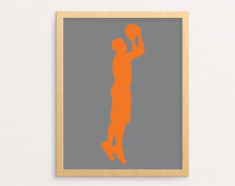 May include: A framed print featuring an orange silhouette of a basketball player mid-shot against a gray background. The artwork is encased in a light brown wooden frame, suitable for sports-themed decor. The player is depicted in a dynamic pose.