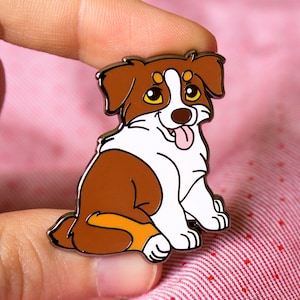 Puede incluir: Un pin de esmalte de perro pastor australiano marrón y blanco con una expresión feliz y la lengua fuera. El pin está sentado sobre un fondo de tela rosa y blanca.