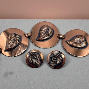 Puede incluir: Un conjunto de joyas de color cobre con un brazalete y pendientes a juego. El brazalete tiene tres eslabones circulares con diseños de hojas, y los pendientes son pequeños, redondos y también presentan diseños de hojas. El conjunto se exhibe sobre una superficie gris.