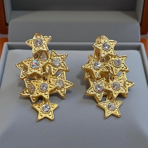 May include: A pair of gold star earrings. Each earring has five stars, each with a central clear gemstone. The earrings are displayed in a grey jewellery box.