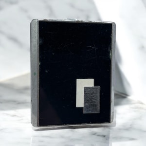 May include: A rectangular, silver-toned metal compact with a black enamel face. The face features a white and gray geometric design. The compact is closed and has a small, silver clasp.