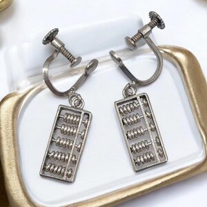 May include: A pair of silver-toned clip-on earrings. Each earring features a rectangular abacus charm with movable beads. The clip-on mechanism has a screw-like design for secure attachment. The earrings are displayed on a white surface.