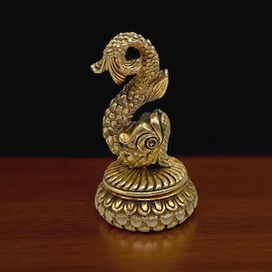 May include: A gold-colored, ornate fish figurine with a detailed, scaled body and a curved tail. The fish sits atop a round, decorative base with a scalloped edge. The background is dark, highlighting the figurine.