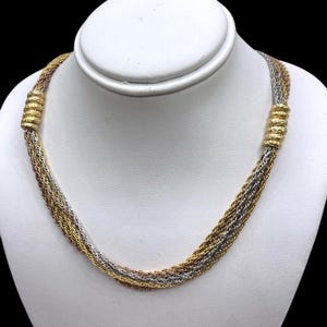 Puede incluir: Un collar de múltiples hilos con tonos dorados, plateados y oro rosa. El collar se exhibe sobre un busto blanco y presenta detalles dorados texturizados en cada extremo. Los hilos están trenzados, creando un diseño único y elegante.