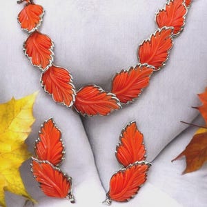 May include: A vintage jewelry set featuring a necklace and matching earrings. The set showcases orange leaf-shaped pendants with silver-colored trim. The jewelry is displayed on a white surface, with autumn leaves in the background.
