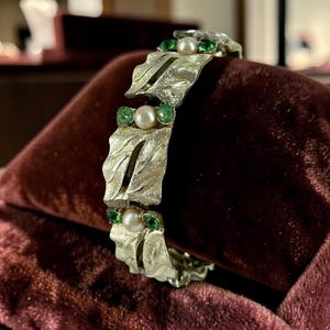May include: A gold-tone metal link bracelet featuring a textured leaf-like design. Each link is accented with a central round white pearl flanked by two small, vibrant green faceted stones, creating a repeating pattern along the entire length.