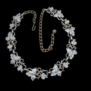 May include: A vintage necklace with a leaf design. The necklace features silver-coloured leaves with a textured surface, connected by gold-toned metal. The necklace has a hook clasp and an adjustable chain.