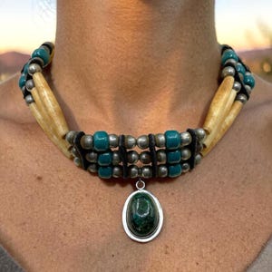Op de afbeelding: Een handgemaakte ketting met een groene stenen hanger in een zilverkleurig frame. De ketting heeft meerdere strengen van botkleurige kralen, turquoise kralen en zilverkleurige kralen, waardoor een gelaagd effect ontstaat. De ketting is ontworpen om om de nek te worden gedragen.