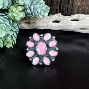 May include: A silver ring featuring a cluster of pink oval-shaped stones surrounding a larger oval center stone. The ring has a detailed, textured silver setting and is displayed on a black surface.