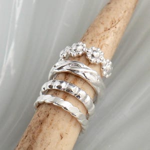 May include: A collection of silver rings with various designs, including floral, twisted, and textured bands. The rings are displayed on a light brown, cylindrical object against a soft, blurred background.