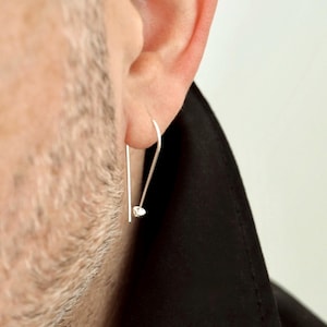 May include: A close-up of a silver earring with a small, clear gemstone. The earring is a simple, linear design, with a hook that curves over the ear. The earring is worn by a person.