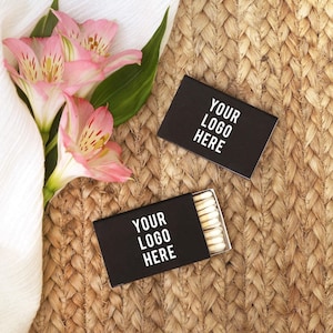Puede incluir: Dos cajas de cerillas rectangulares negras con el texto blanco YOUR LOGO HERE impreso en la parte frontal. Una caja está cerrada y la otra está parcialmente abierta, revelando una fila de cerillas de madera con punta blanca. Flores de alstroemeria rosas y blancas.