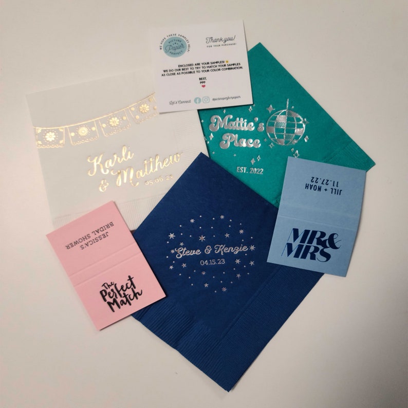 May include: A collection of wedding-themed napkins with various designs. The napkins are blue, teal, pink, and light blue. The designs include a disco ball, a star with the text "Steve & Kenzie" and the date "04.15.23", a banner with the text "Karli & Matthew" and the date "05.00.24", and a banner with the text "MR & MRS".