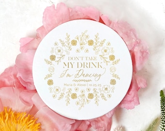 Könnte beinhalten: Ein runder weißer Untersetzer mit einem goldenen Blumenkranz-Design, das den Text "DON'T TAKE MY DRINK I'm Dancing" sowie die Namen "Maria & Alexei" und das Datum "25.10.25" in goldener Schrift in der Mitte umschließt.