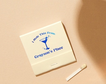 Puede incluir: Una caja de cerillas de color crema con un gráfico azul de una copa de martini con una aceituna y el texto "I Stole This From: Grayson's Place" impreso en tinta azul. Una cerilla de madera se encuentra junto a la caja sobre una superficie.