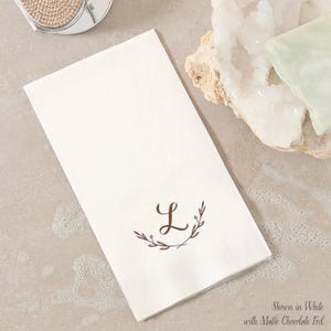 May include: A rectangular white paper napkin featuring a brown printed letter L centered within a delicate botanical wreath design. The text at the bottom right corner reads Shown in White with Matte Chocolate Foil, indicating the product color.