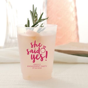 May include: A clear plastic cup with a pink design that says "She said yes!" with a ring and diamond graphic. The text below says "Emily's Engagement Party 07.24.2023".