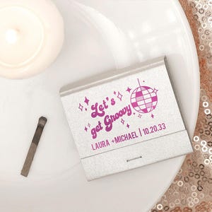 Puede incluir: Una caja de cerillas plateada con un gráfico rosa de una bola de discoteca y el texto "Let's get Groovy", los nombres "LAURA + MICHAEL" y la fecha "10.20.33". Una cerilla usada se encuentra al lado sobre una superficie blanca.