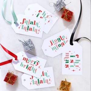 Puede incluir: Una colección de etiquetas de regalo rectangulares blancas con texto festivo en rojo y verde que incluye 'fa la la', 'jingle and mingle', 'merry and bright' y 'christmas', decoradas con estrellas y acompañadas de cajas de regalo en miniatura.