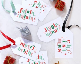 Puede incluir: Una colección de etiquetas de regalo rectangulares blancas con texto festivo en rojo y verde que incluye 'fa la la', 'jingle and mingle', 'merry and bright' y 'christmas', decoradas con estrellas y acompañadas de cajas de regalo en miniatura.