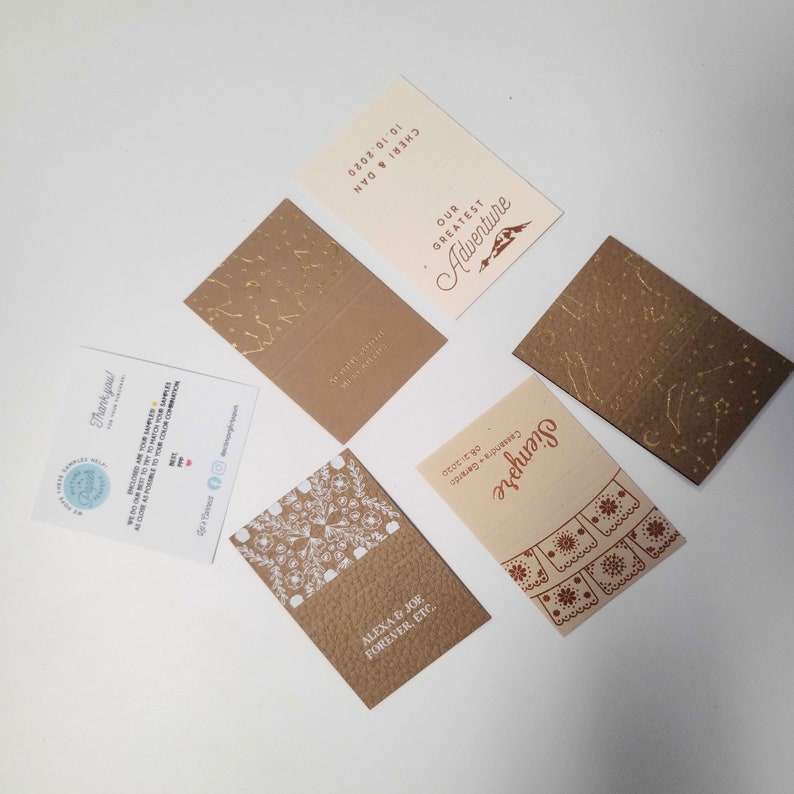 May include: A set of six wedding invitation cards with different designs. The cards are printed on brown paper with gold foil accents. The text on the cards includes the names of the couple, the date of the wedding, and the phrase "Our Greatest Adventure".