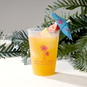 Puede incluir: Un vaso de plástico translúcido lleno de una bebida naranja, hielo y trozos de fruta. El vaso presenta un gráfico rosa de dos palmeras y el texto JENN NEST & BESTS ALBANY, BAHAMAS | 2023. Una pequeña sombrilla de cóctel azul está insertada.