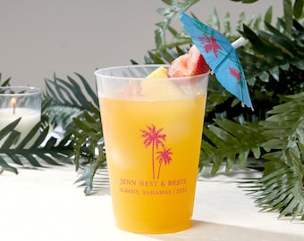 Puede incluir: Un vaso de plástico translúcido lleno de una bebida naranja, hielo y trozos de fruta. El vaso presenta un gráfico rosa de dos palmeras y el texto JENN NEST & BESTS ALBANY, BAHAMAS | 2023. Una pequeña sombrilla de cóctel azul está insertada.