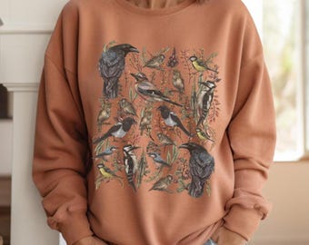 May include: A rust-colored sweatshirt featuring a detailed illustration of various birds, including crows, magpies, and smaller songbirds. The design is set against a backdrop of foliage. The sweatshirt is a crew neck style and is made of a soft material.