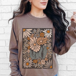 May include: A long-sleeved brown shirt featuring a floral design. The graphic includes large daisies in shades of peach, cream, and orange, surrounded by green leaves and smaller flowers. The shirt has a crew neck and a relaxed fit.
