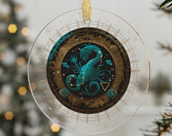 May include: A round glass ornament with a gold rim and a black background. The ornament features a blue and gold illustration of the Scorpio zodiac sign with the symbol "m" on either side of the scorpion. The background is a dark blue with gold stars.