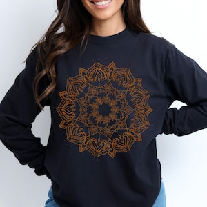 May include: Black long-sleeve shirt featuring a detailed, golden mandala design on the front. The intricate pattern is centered and symmetrical, with heart-shaped elements. The shirt is a solid color, with a crew neck and long sleeves.