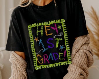 May include: A black short-sleeve t-shirt featuring a neon green rectangular border with a chalk-like texture. Inside the border, the text reads "HEY 1ST GRADE!" in colorful, textured lettering, accented by small star and arrow graphic elements.