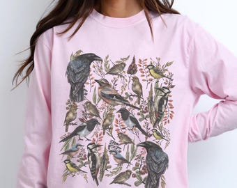 May include: A light pink long-sleeve shirt featuring a detailed graphic print of various birds, including ravens, woodpeckers, and smaller songbirds, interspersed with botanical elements like leaves and branches in muted earth tones.