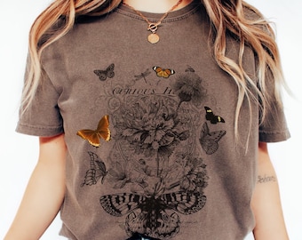 May include: A brown t-shirt featuring a detailed butterfly and floral design. The graphic includes several butterflies in shades of brown and orange, with the text "Curious Life" in a decorative font. The shirt is a neutral color and has short sleeves.