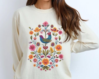May include: Cream-colored long-sleeve sweatshirt featuring a colorful floral and bird design. The design includes a blue bird, pink, orange, and yellow flowers, and green leaves arranged in a circular pattern. The sweatshirt is made of a soft material.