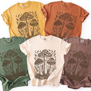 May include: Five short-sleeve crew neck t-shirts in yellow, rust orange, sage green, cream, and brown. Each shirt features a dark brown graphic print of three mushrooms surrounded by ferns and foliage centered on the front of the garment.