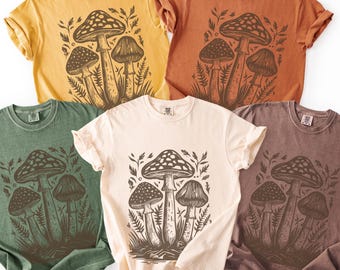 May include: Five short-sleeve crew neck t-shirts in yellow, rust orange, sage green, cream, and brown. Each shirt features a dark brown graphic print of three mushrooms surrounded by ferns and foliage centered on the front of the garment.