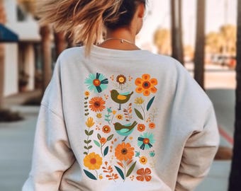 May include: A light beige long-sleeve sweatshirt featuring a large rectangular graphic on the back. The print displays a collection of colorful flowers in shades of orange, yellow, and teal, interspersed with two small birds in green and teal tones.