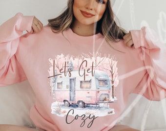 May include: A light pink sweatshirt with a graphic of a vintage camper in a winter scene. The text "Let's Get Cozy" is printed above and below the camper. The sweatshirt is a soft, comfortable material.