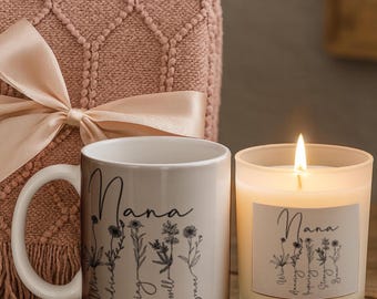 May include: A white ceramic mug and a frosted glass candle, both featuring the word Nana in black script above a line of black wildflower illustrations and descriptive words. The mug and candle are positioned in front of a folded dusty pink knit blanket.