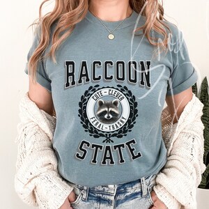 May include: A blue t-shirt with a graphic of a raccoon in a circle with the words "Cute - Clever" above the raccoon and "Feral - Trashy" below the raccoon. The words "Raccoon State" are printed below the circle.