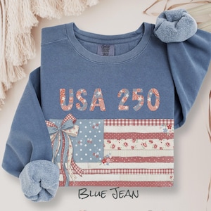 Puede incluir: Sudadera azul con el texto "USA 250" y un diseño de bandera americana de patchwork. La sudadera tiene cuello redondo y mangas largas con una scrunchie a juego. El texto "Blue Jean" está en la parte inferior.